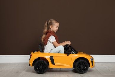 Photo of Cute little girl driving children's electric toy car near brown wall indoors