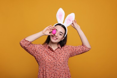 Photo of Happy woman in bunny ears headband holding painted Easter egg on orange background