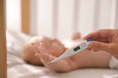 Photo of Cute baby lying in crib, focus on woman holding digital thermometer. Health care