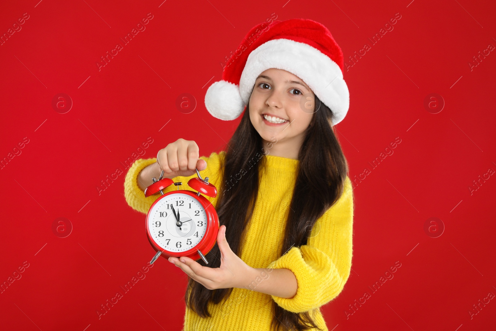 Photo of Girl in Santa hat with alarm clock on red background. Christmas countdown