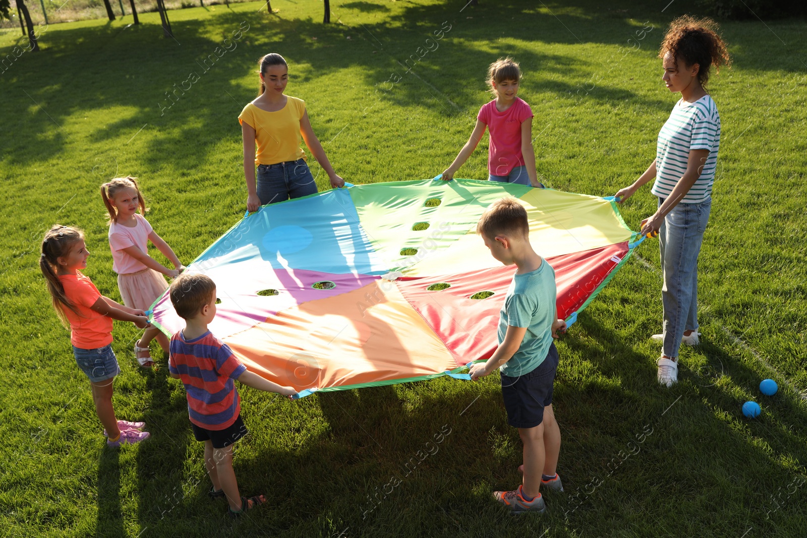 Photo of Group of children and teachers playing with rainbow playground parachute on green grass. Summer camp activity