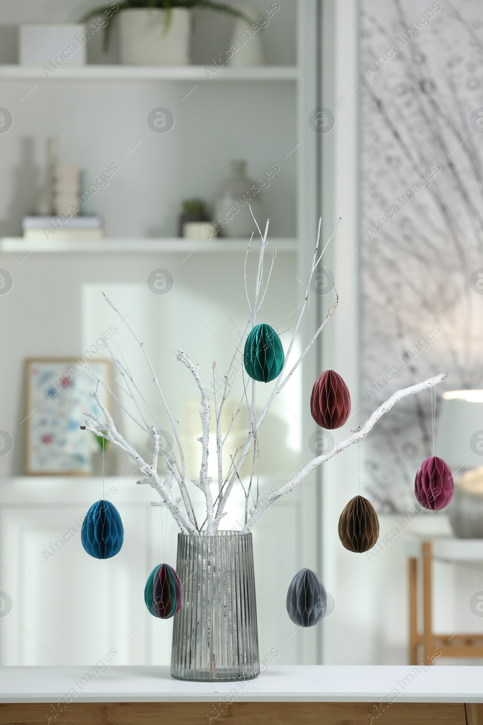 Photo of Branches with paper eggs in vase on white table at home. Beautiful Easter decor