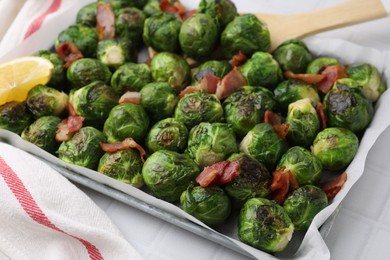 Delicious roasted Brussels sprouts, bacon and lemon in baking dish on white tiled table, closeup