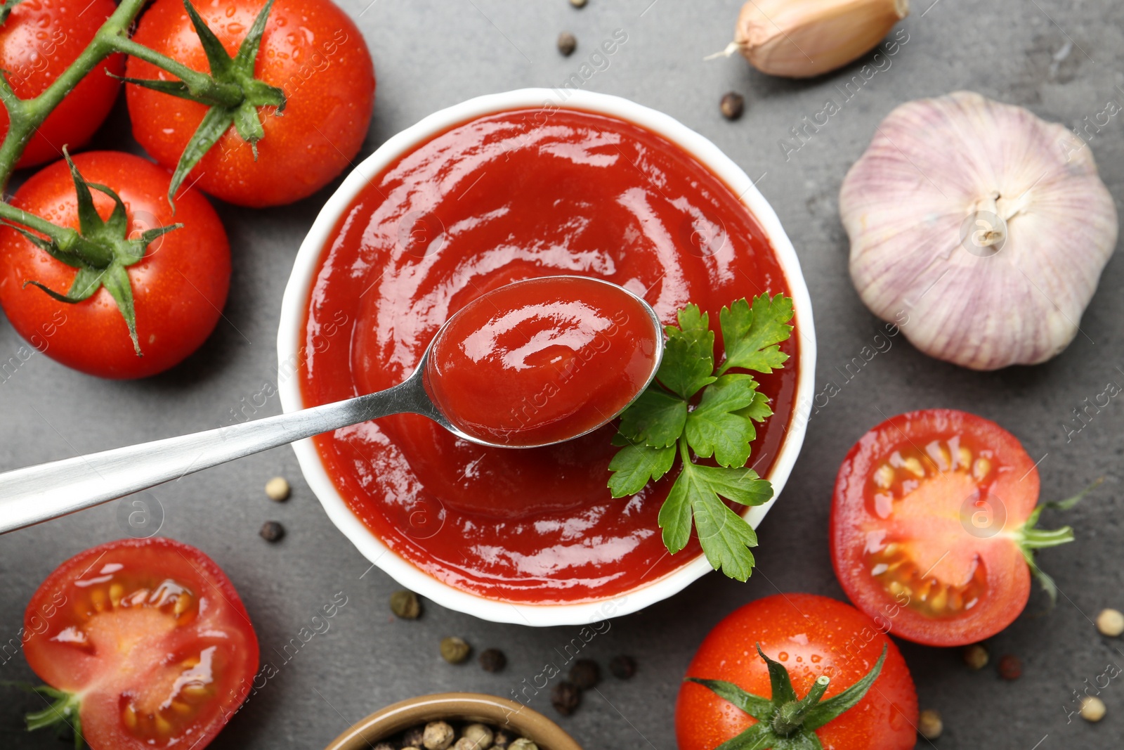 Photo of Taking delicious tomato ketchup with spoon from bowl at grey textured table with spices and products, flat lay