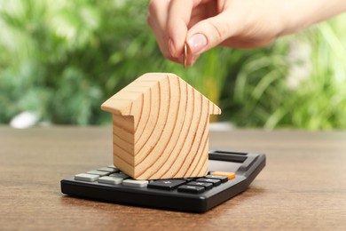 Mortgage concept. Woman putting coin into house model at wooden table against blurred green background, closeup