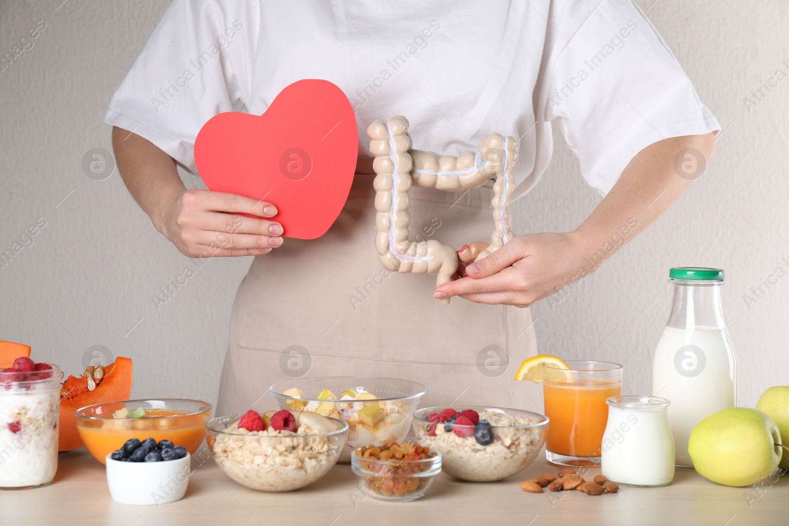 Photo of Woman holding paper heart and large intestine model near table with food, closeup. Balanced nutrition for healthy digestive system