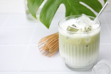 Photo of Glass of tasty iced matcha latte, bamboo whisk and leaf on white tiled table, closeup. Space for text