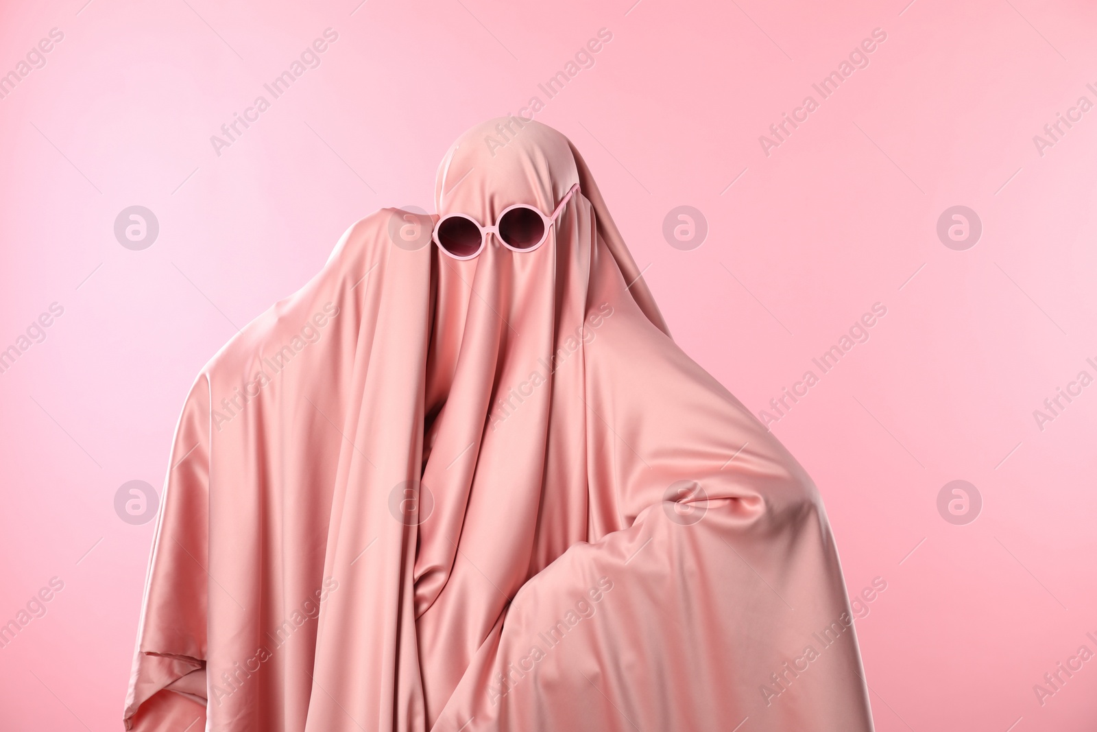 Photo of Glamorous ghost. Woman in sheet with sunglasses on pink background, space for text