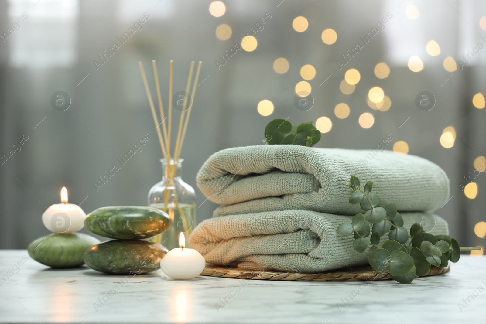 Photo of Spa composition. Towels, stones, reed air freshener and burning candles on white marble table against blurred lights