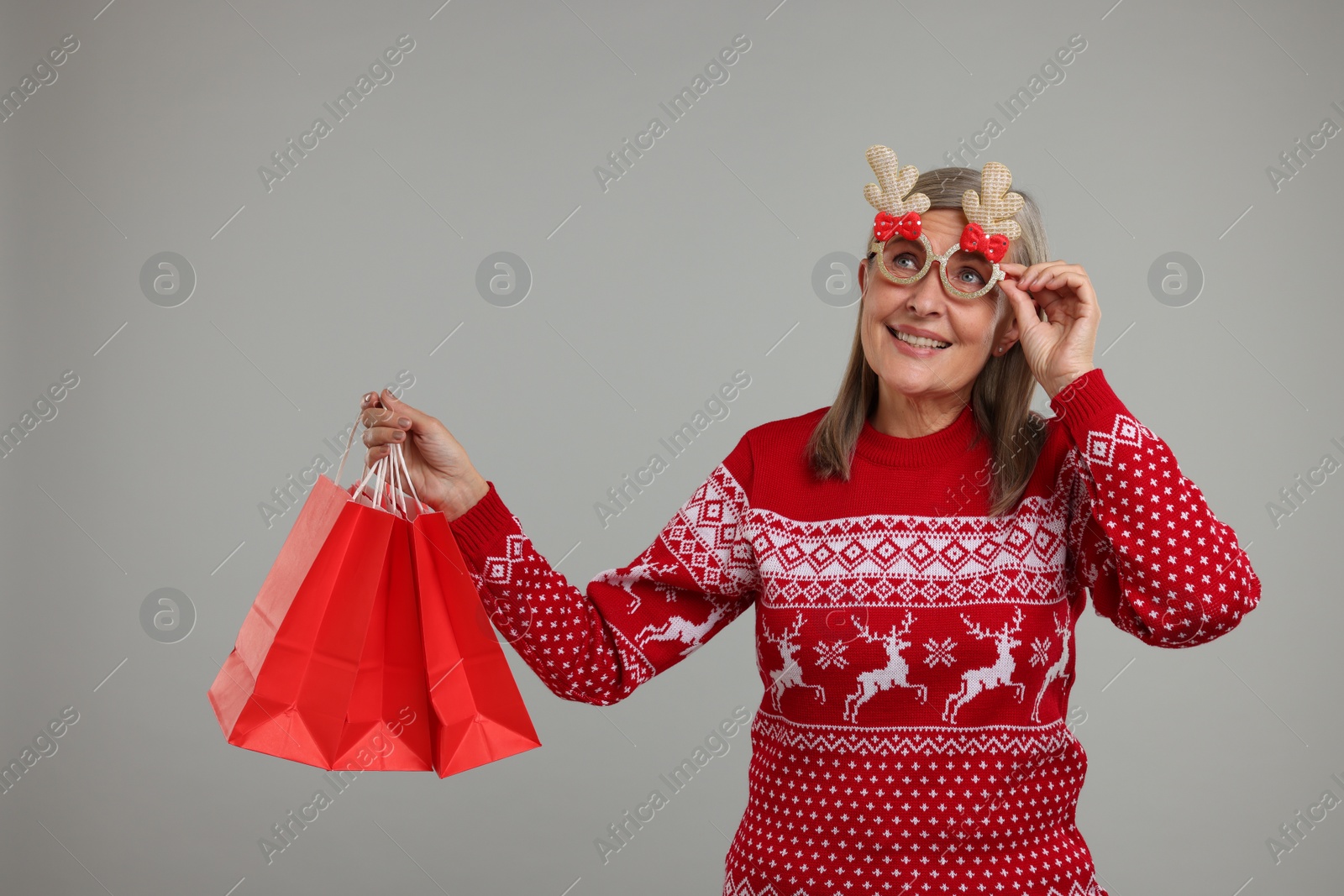 Photo of Happy senior woman in Christmas sweater and funny glasses with shopping bags on grey background