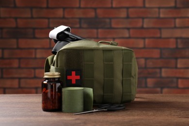 Military first aid kit, tourniquet, pills and elastic bandage on wooden table against brick wall