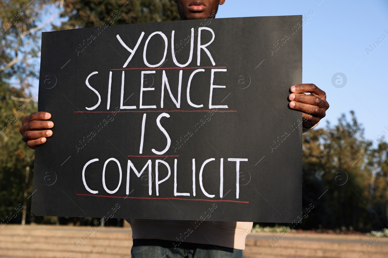 Photo of African American man holding sign with phrase Your Silence Is Complicit outdoors, closeup. Racism concept