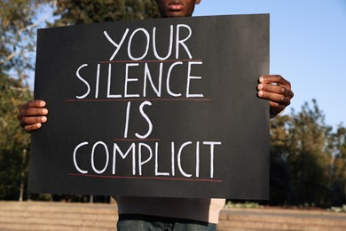 Photo of African American man holding sign with phrase Your Silence Is Complicit outdoors, closeup. Racism concept