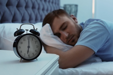 Photo of Alarm clock on table and young man sleeping in bed at night