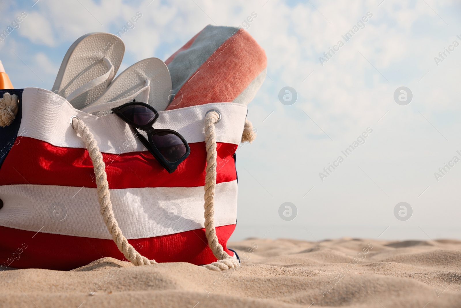 Photo of Beach bag with flip flops, towel and sunglasses on sand, space for text