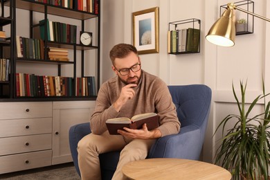 Photo of Young man reading book in armchair indoors. Home library