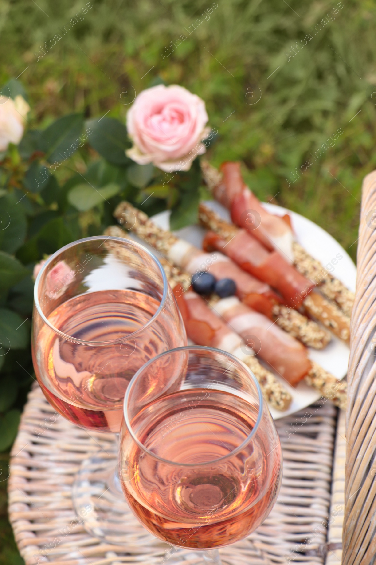 Photo of Flowers near glasses of delicious rose wine and food on picnic basket outdoors, closeup