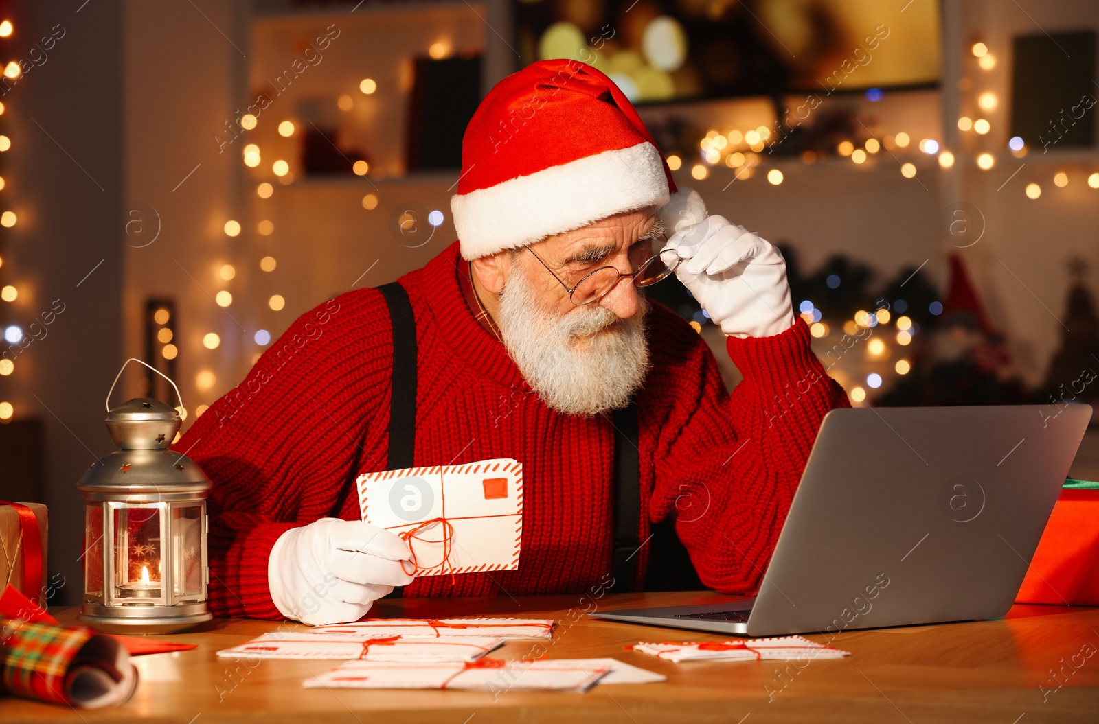 Photo of Santa Claus using laptop and holding letter at his workplace in room decorated for Christmas