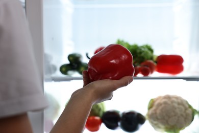 Young woman taking red bell pepper out of refrigerator at night, closeup