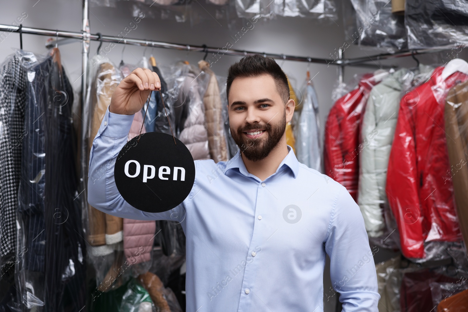 Photo of Dry-cleaning service. Happy worker holding Open sign near clothes in plastic bags indoors