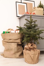 Beautiful fir tree and sack with Christmas gifts near chest of drawers indoors