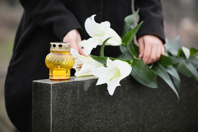 Woman holding white lilies near black granite tombstone with candle outdoors, closeup. Funeral ceremony