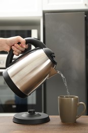 Photo of Woman pouring hot water from electric kettle into cup in kitchen, closeup