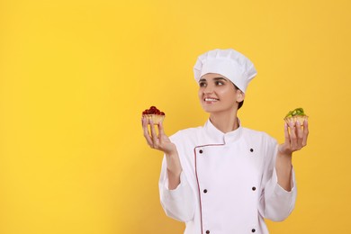 Photo of Happy professional confectioner in uniform with delicious tartlets on yellow background. Space for text