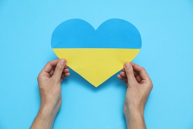 Photo of Man holding paper heart in colors of Ukrainian flag on light blue background, top view