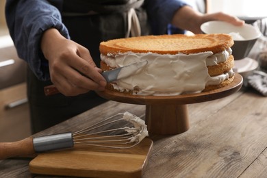 Photo of Woman smearing sides of sponge cake with cream at wooden table, closeup