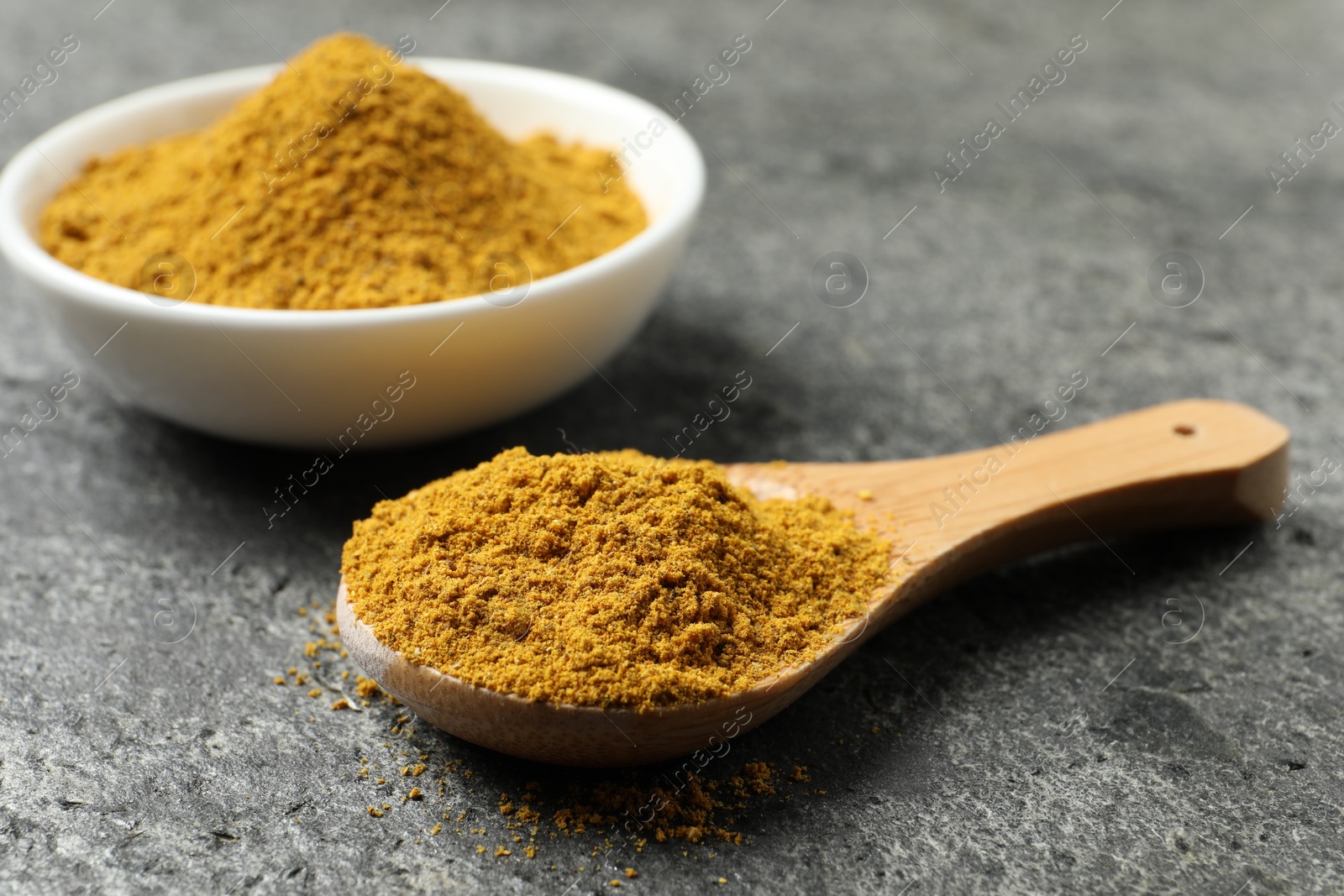 Photo of Spoon and bowl with dry curry powder on dark textured table, closeup
