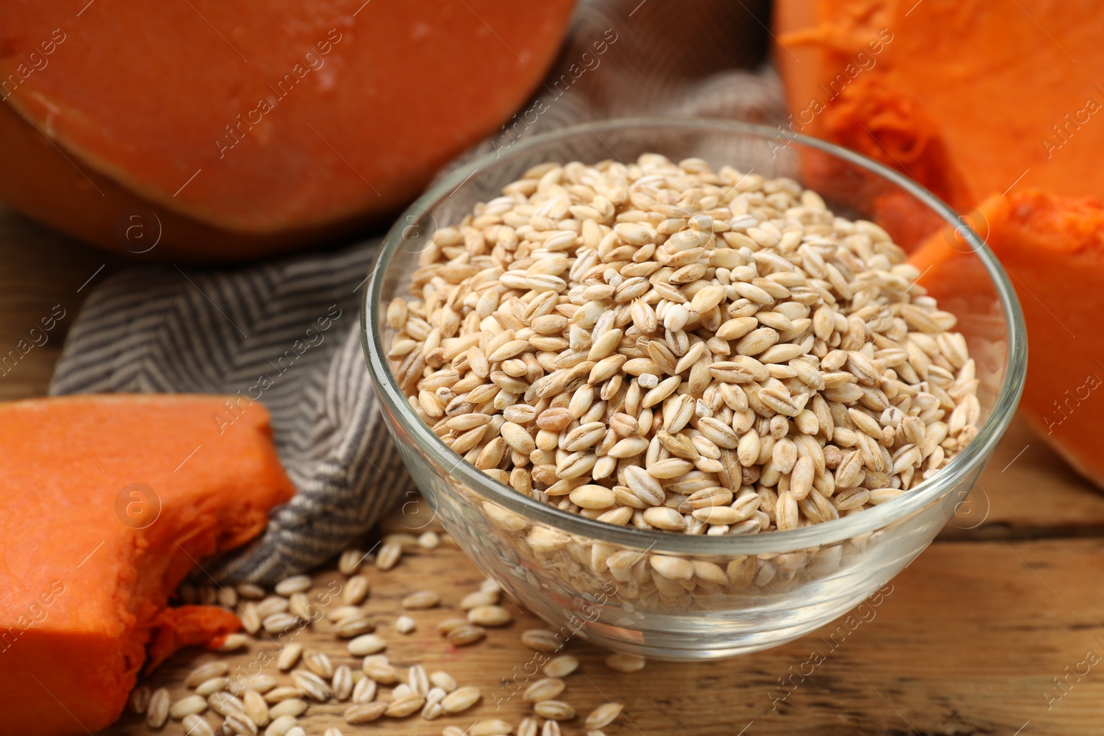 Photo of Dry pearl barley in bowl and pieces of pumpkin on wooden table, closeup