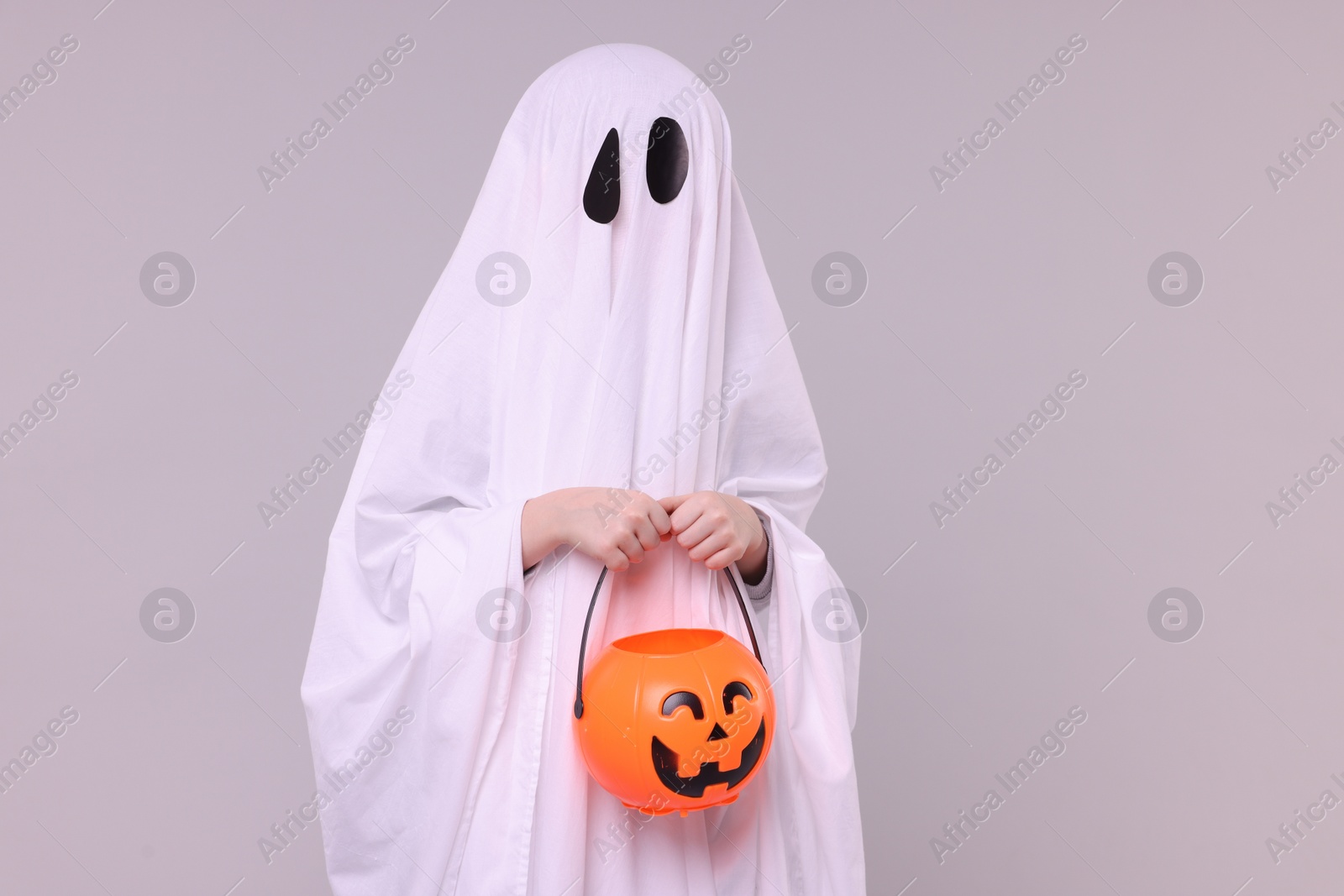 Photo of Child in white ghost costume holding pumpkin bucket on light grey background. Halloween celebration