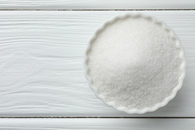 Photo of Granulated sugar in bowl on white wooden table, top view. Space for text