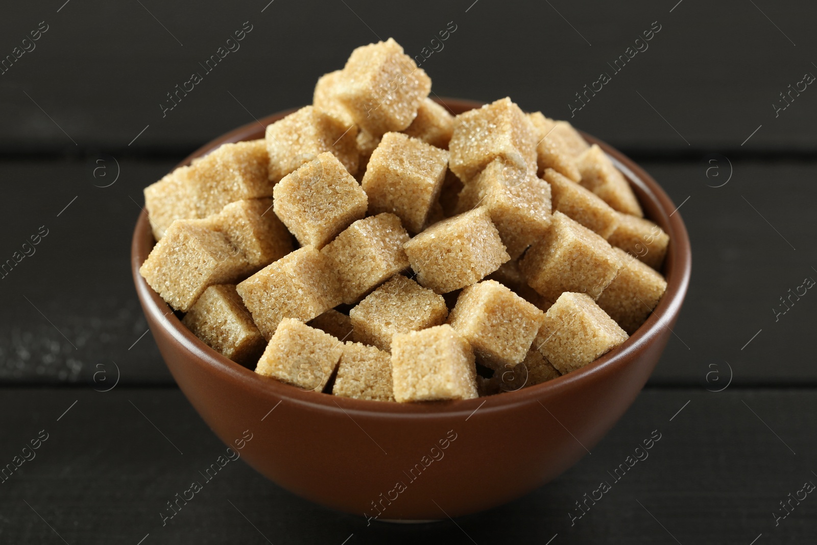 Photo of Brown sugar cubes in bowl on black wooden table, closeup