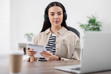 Photo of Young woman in headphones watching webinar at table in room