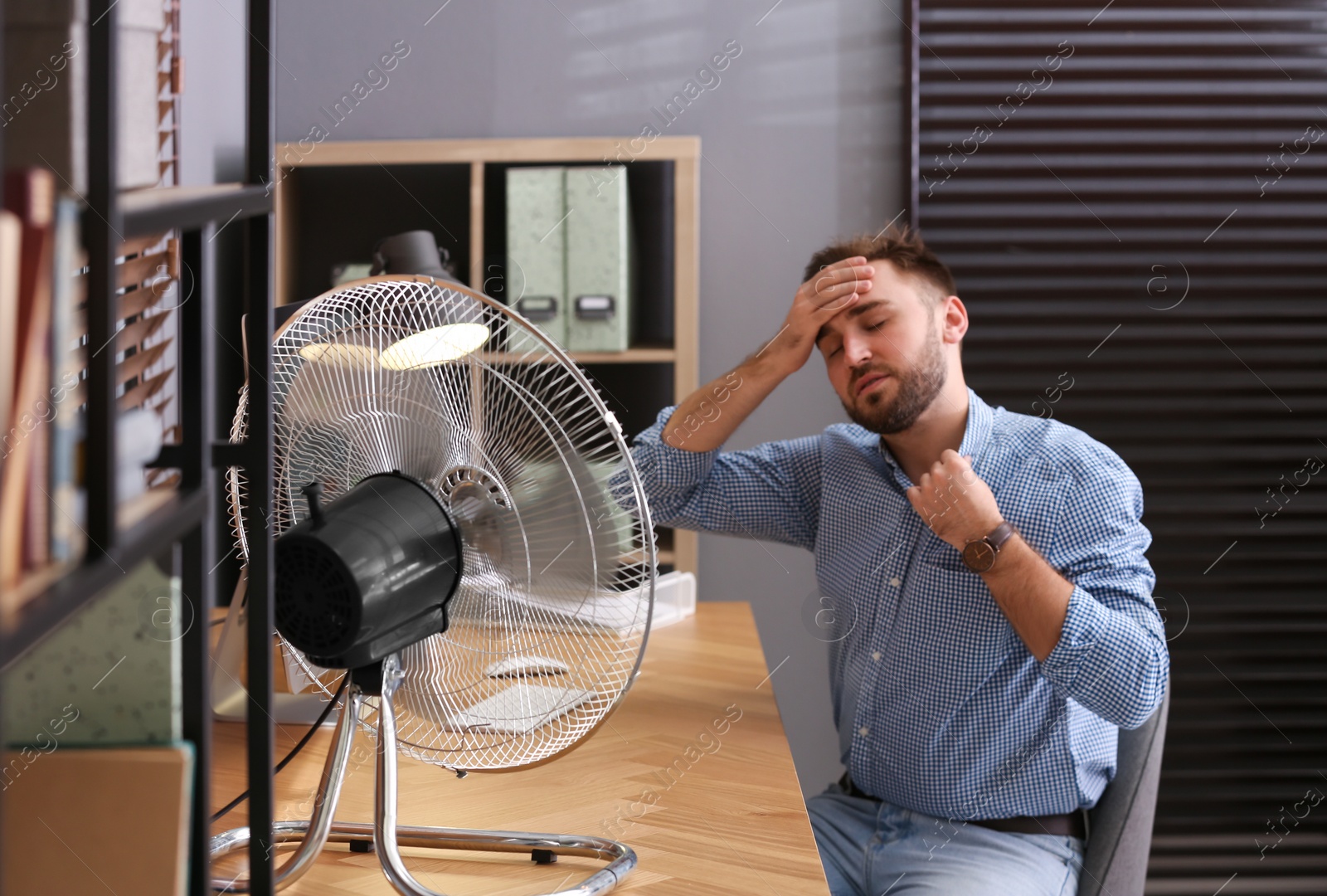 Photo of Man suffering from heat in front of fan at workplace