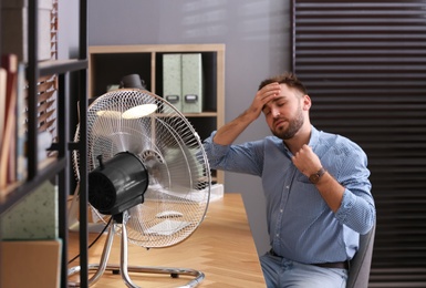 Photo of Man suffering from heat in front of fan at workplace