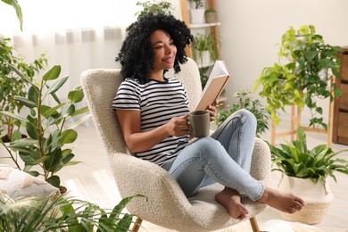 Relaxing atmosphere. Happy woman with cup of hot drink and book sitting in armchair surrounded by houseplants at home