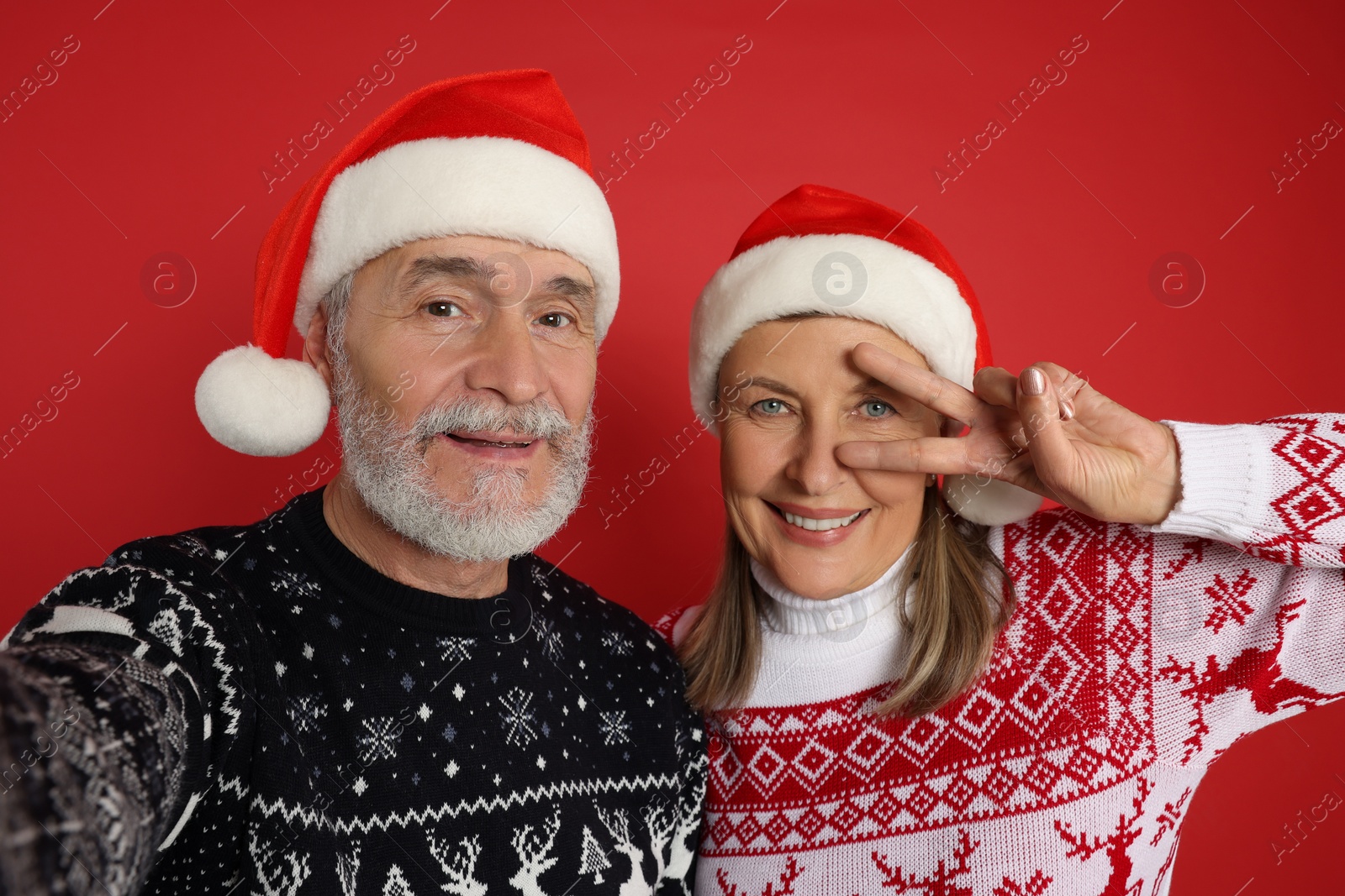 Photo of Senior couple in Christmas sweaters and Santa hats taking selfie on red background