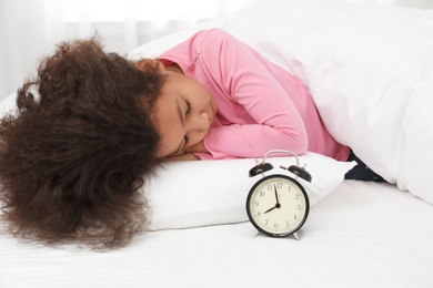 Photo of Cute little African-American girl with alarm clock sleeping in bed