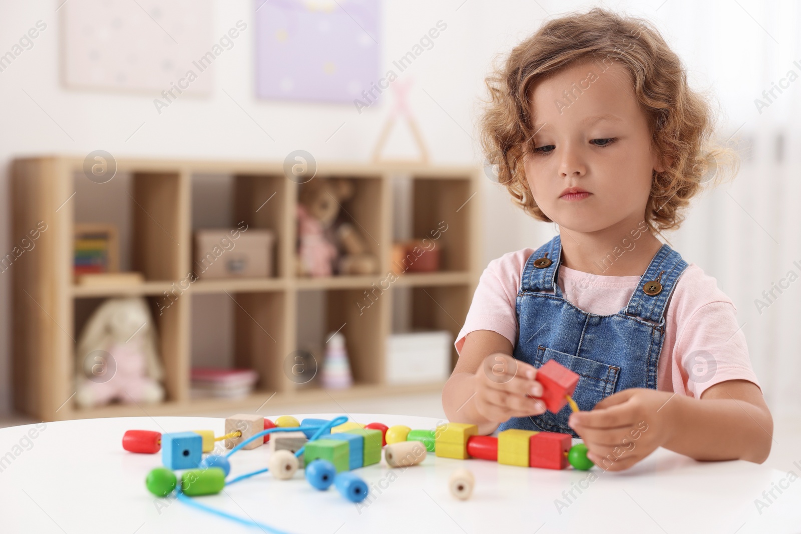 Photo of Motor skills development. Little girl playing with wooden pieces and string for threading activity at table indoors