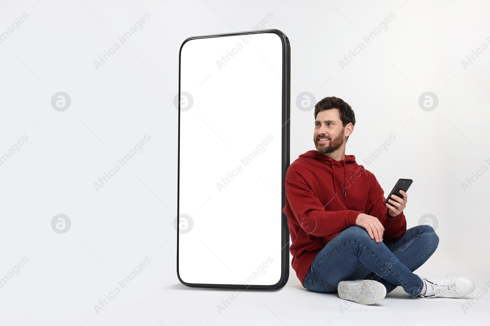 Image of Man with mobile phone sitting near huge device with empty screen on white background. Mockup for design