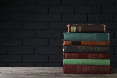 Stack of old hardcover books on wooden table near black brick wall, space for text