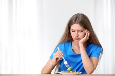 Photo of Portrait of unhappy woman eating broccoli salad at table on light background