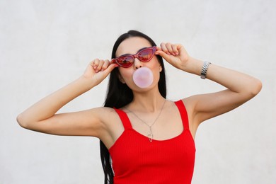 Beautiful woman in stylish sunglasses blowing gum near wall outdoors