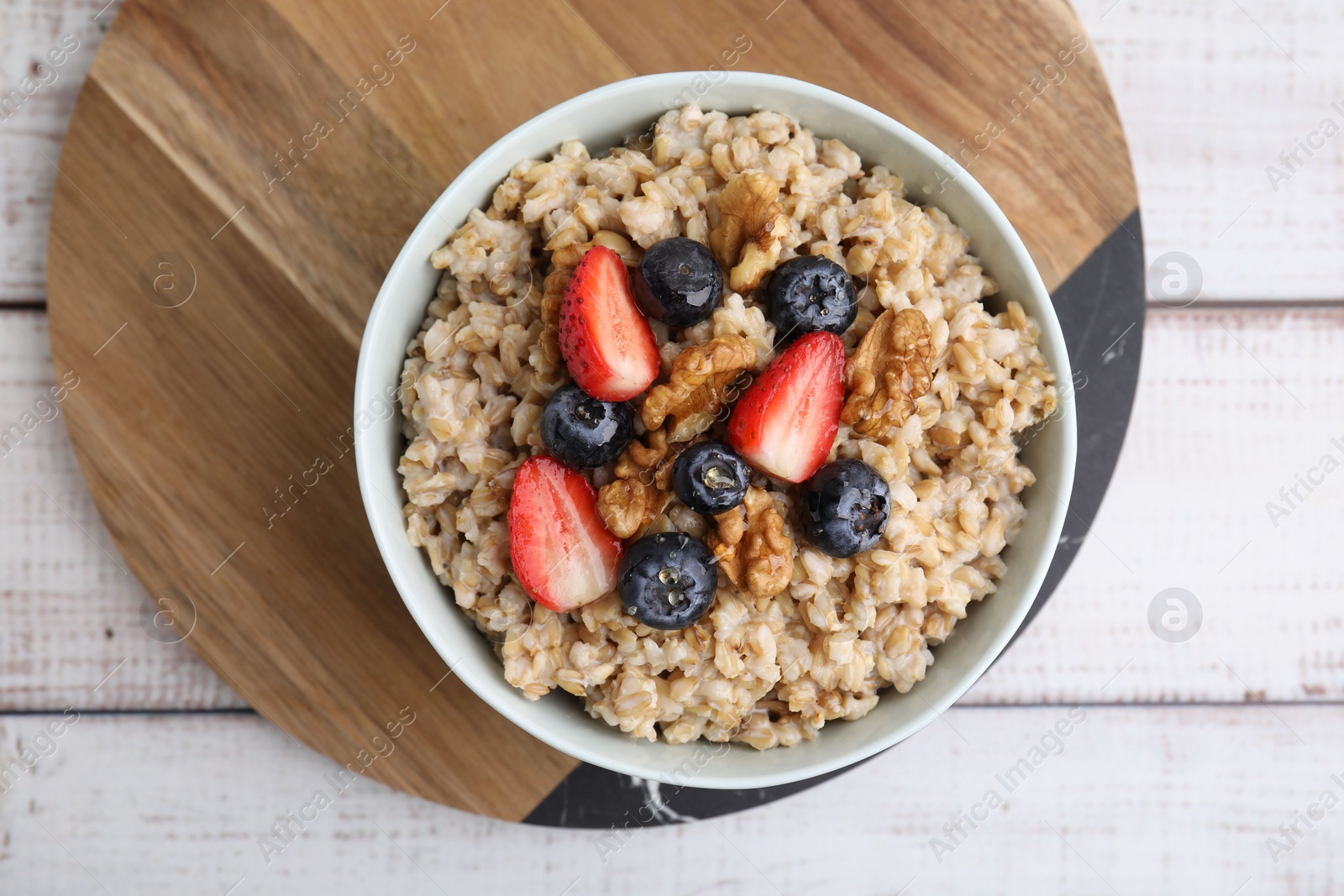 Photo of Tasty oatmeal with strawberries, blueberries and walnuts in bowl on white wooden table, top view