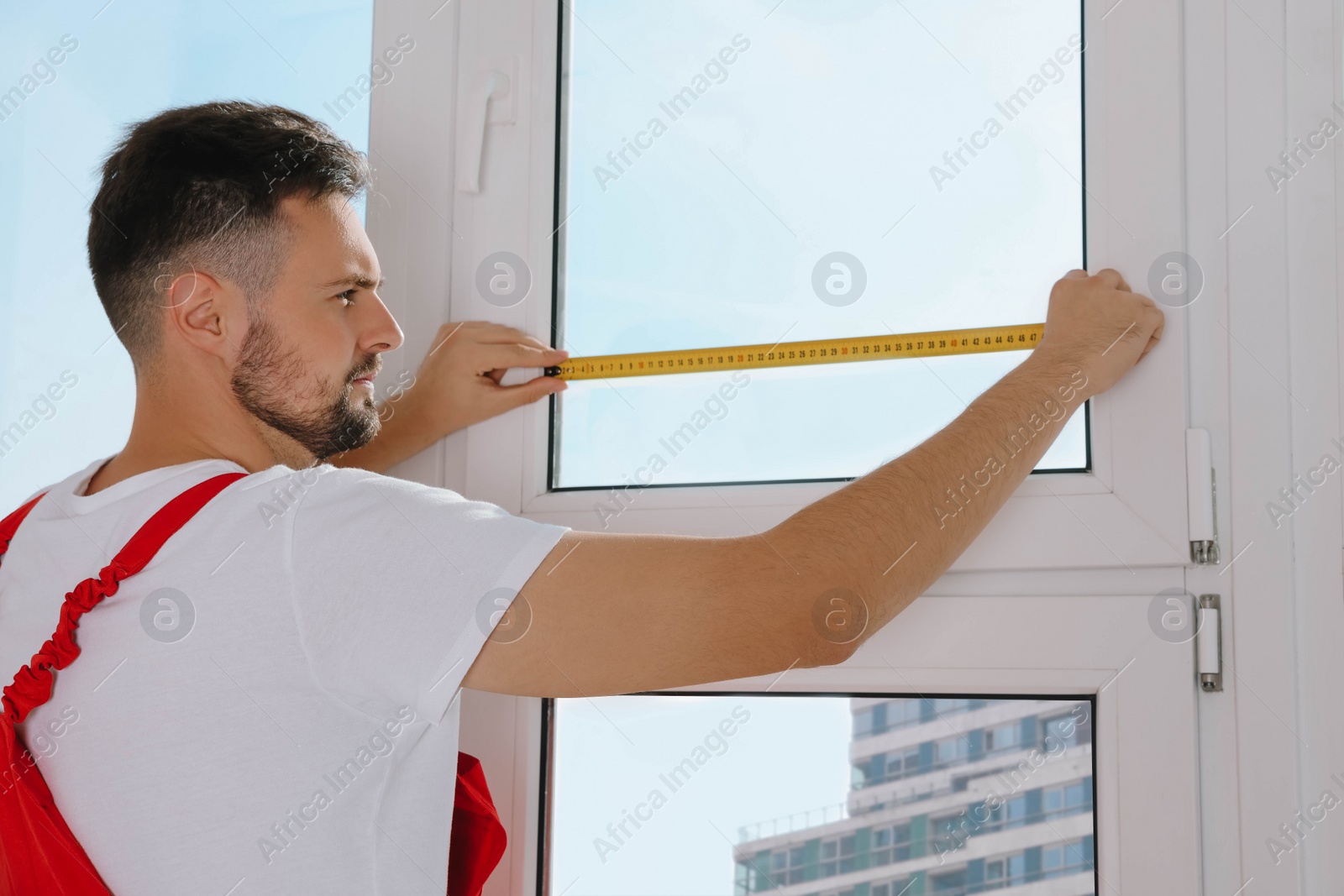 Photo of Worker in uniform measuring window with tape indoors. Roller blinds installation