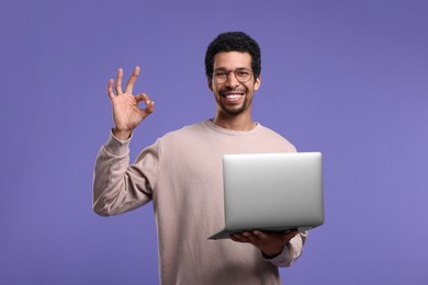 Smiling man with laptop showing ok gesture on purple background