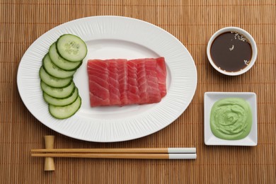 Photo of Tasty sashimi (pieces of fresh raw tuna) served with cucumber slices, soy sauce and wasabi on sushi mat, flat lay